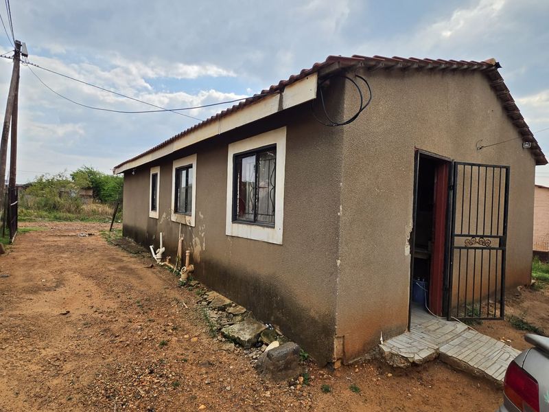 Rear view of a detached house showing exterior walls with visible wear, security gate, and exposed plumbing fixtures on a dirt lot.