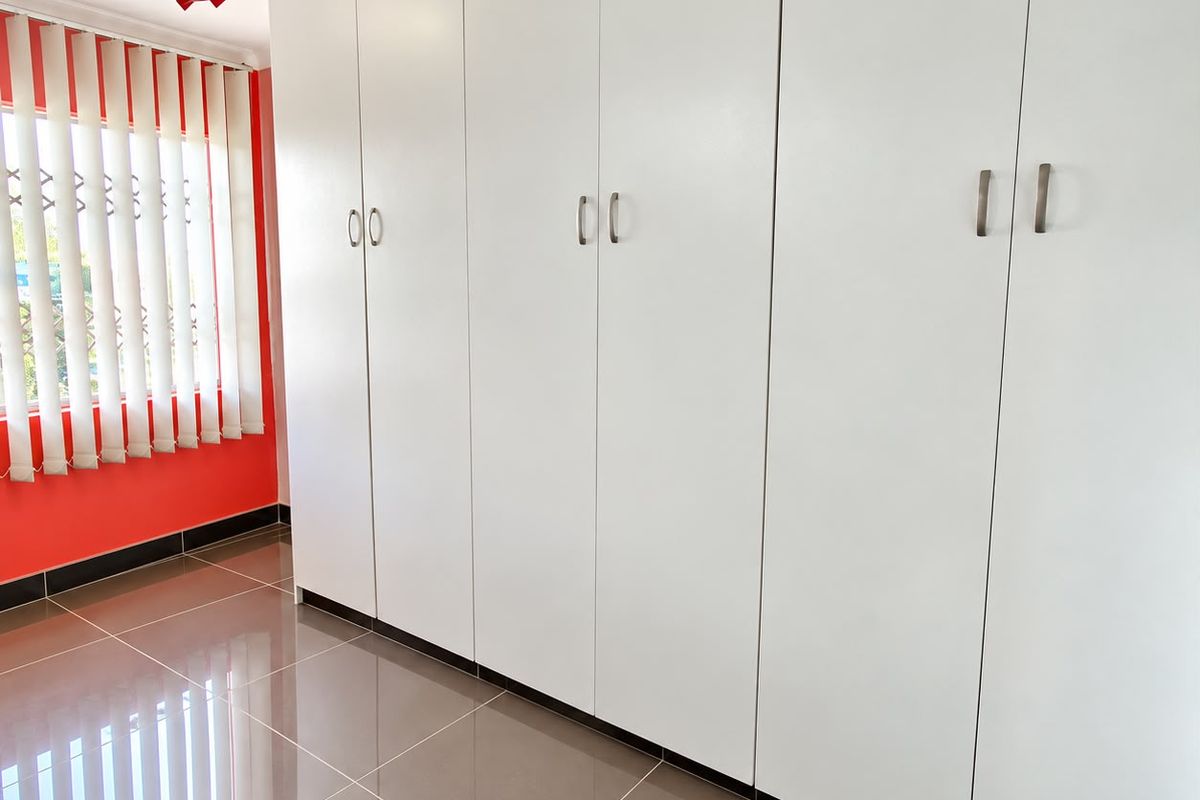 A clean bedroom featuring extensive white built-in wardrobes, neutral tiled flooring, and vertical window blinds.