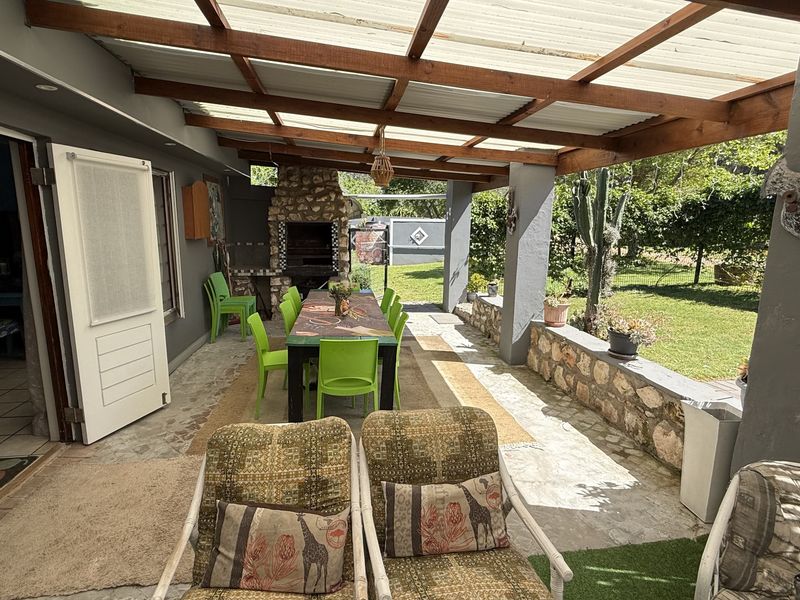 Covered outdoor patio featuring a timber pergola, built-in stone braai, and stone masonry wall, overlooking a private garden area.