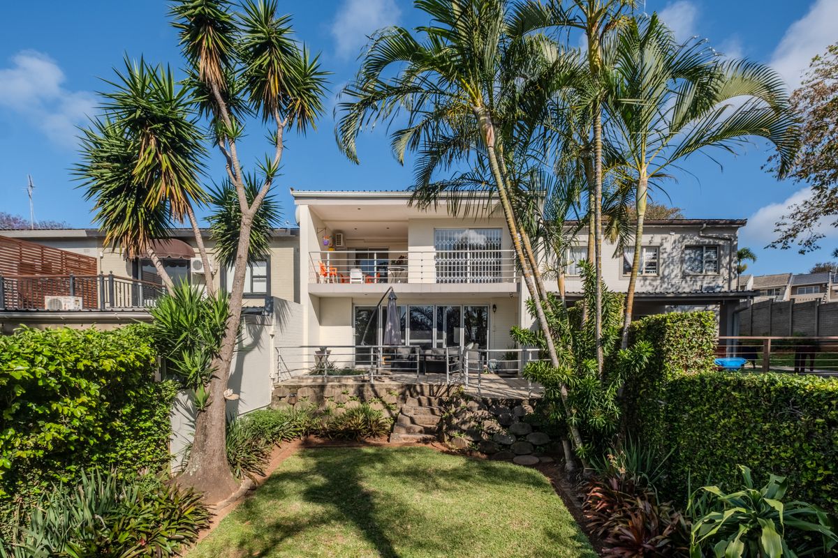 Rear view of the townhouse featuring a landscaped garden, stone retaining wall, ground floor patio, and an upper-level balcony with metal railings.