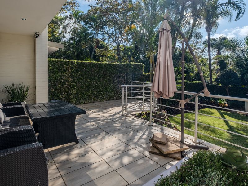 A covered patio area featuring tiled flooring and metal railings, overlooking a private garden with mature landscaping.