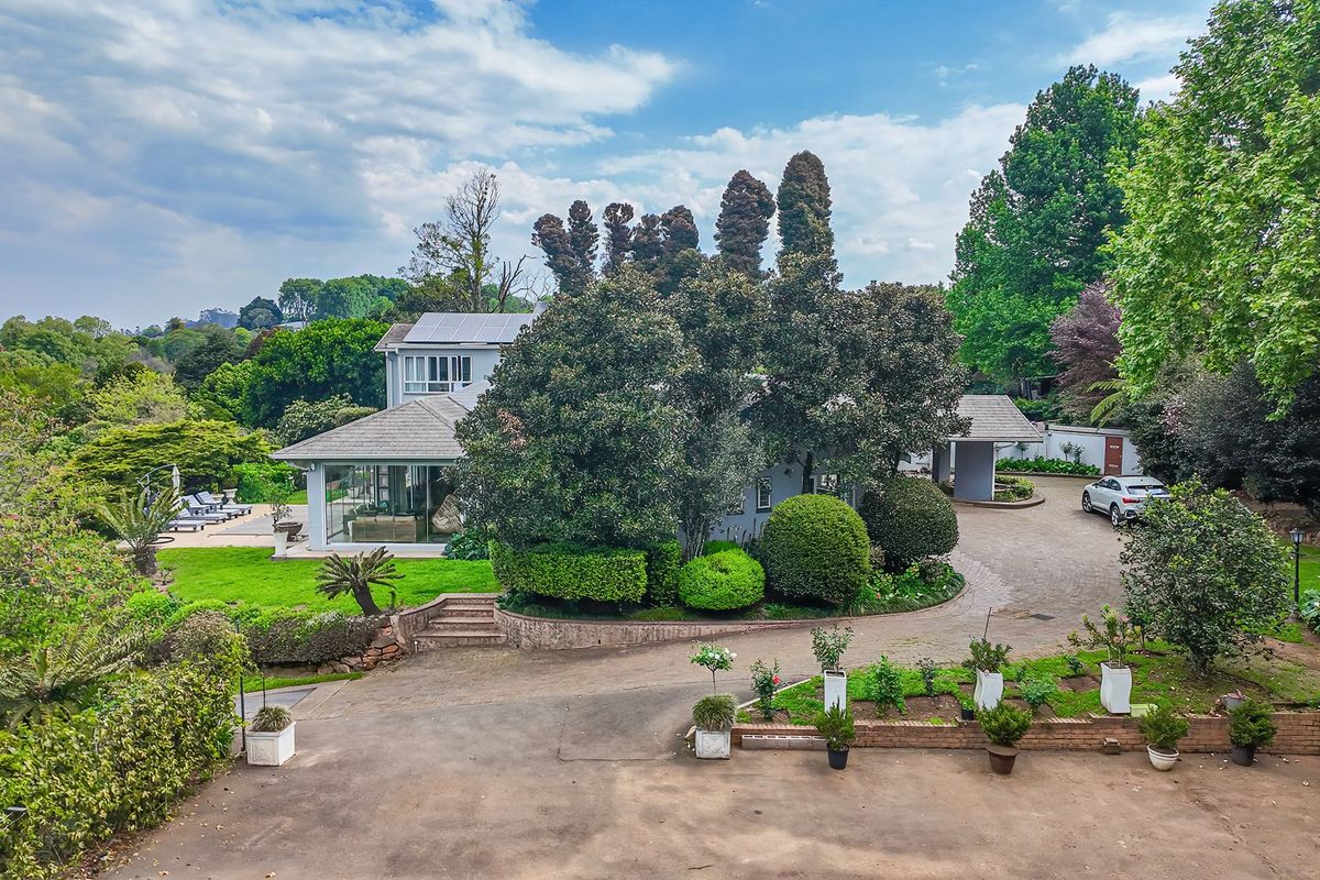 Aerial view of a spacious residential property featuring a large paved driveway, established landscaped gardens, and a main dwelling with solar panels.