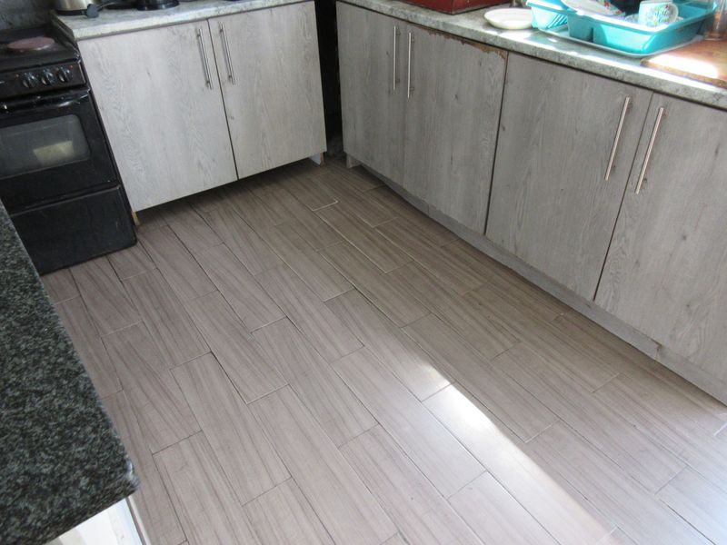 A kitchen area featuring wood-look cabinetry, stone countertops, and tiled flooring, showing signs of wear on the cabinet edges and floor installation.