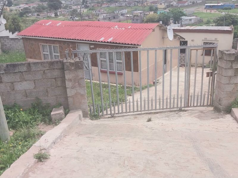 Front view of a single-story house featuring a metal security gate, concrete driveway, and a corrugated metal roof, set within a residential neighborhood.