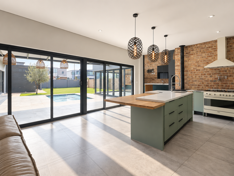 Spacious open-plan kitchen and living area featuring an exposed brick wall, modern kitchen island with breakfast bar, and large sliding doors opening to the pool.