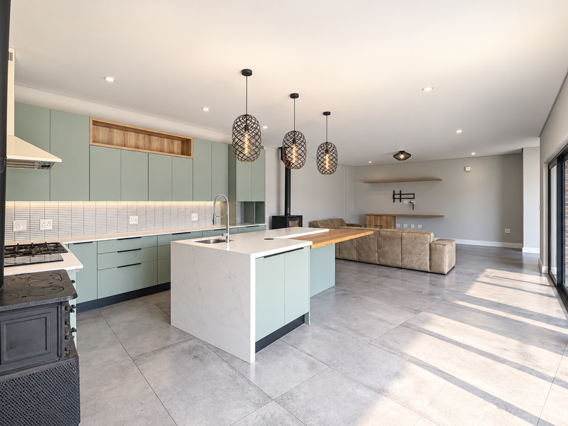 Modern open-plan living area featuring a contemporary kitchen with sage cabinetry, quartz island, tiled floors, and a wood-burning fireplace, opening to outdoor space.