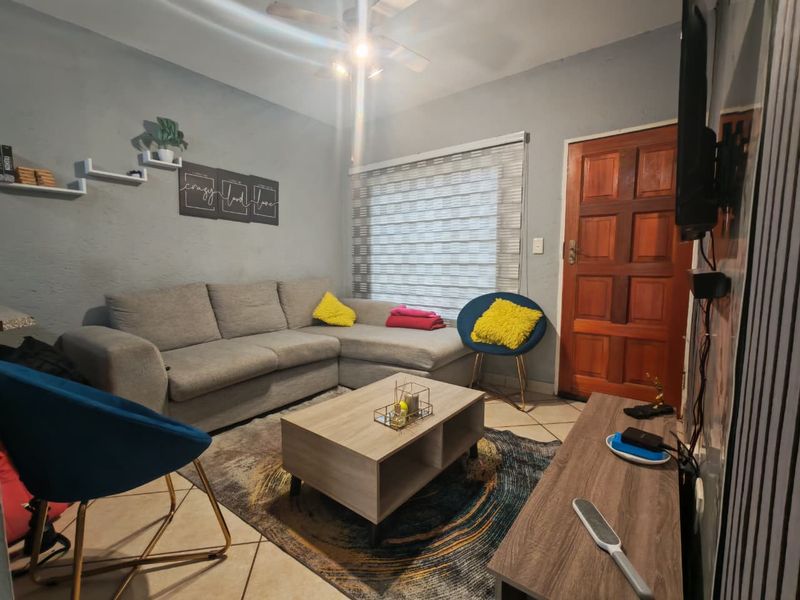 A cozy living room featuring tiled flooring, a ceiling fan, and a wooden entrance door with roller blinds on the window.