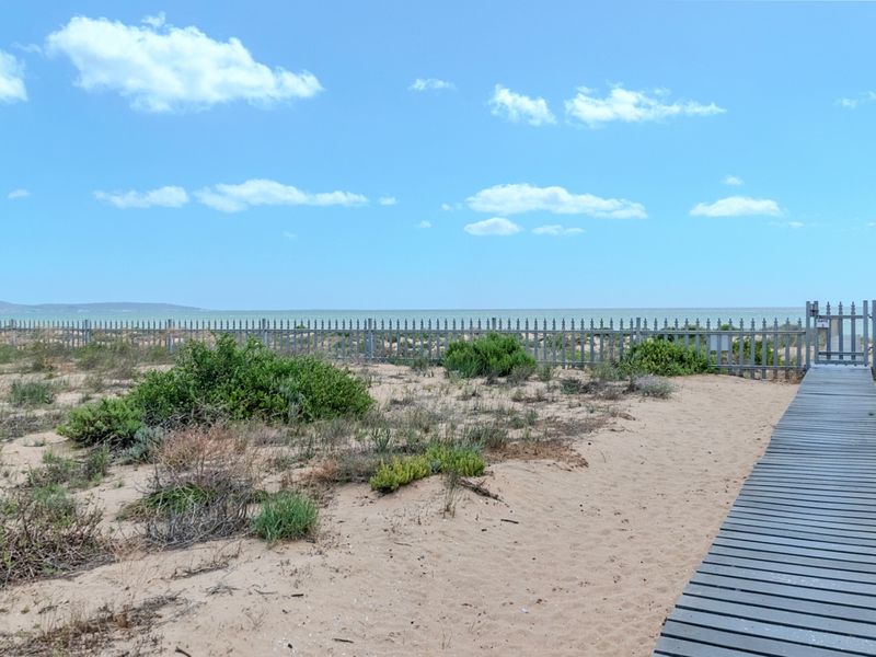 A scenic coastal view featuring a timber boardwalk leading towards a metal fence with direct access to the beach and ocean.
