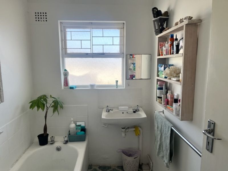 A functional bathroom featuring a wall-mounted basin, a standard bathtub, and a window with security bars, offering a basic and practical layout.