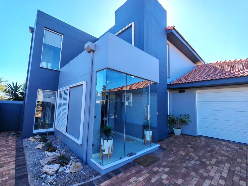 Modern front facade featuring a striking blue exterior, a unique glass-enclosed entrance, paved driveway, and a secure garage.