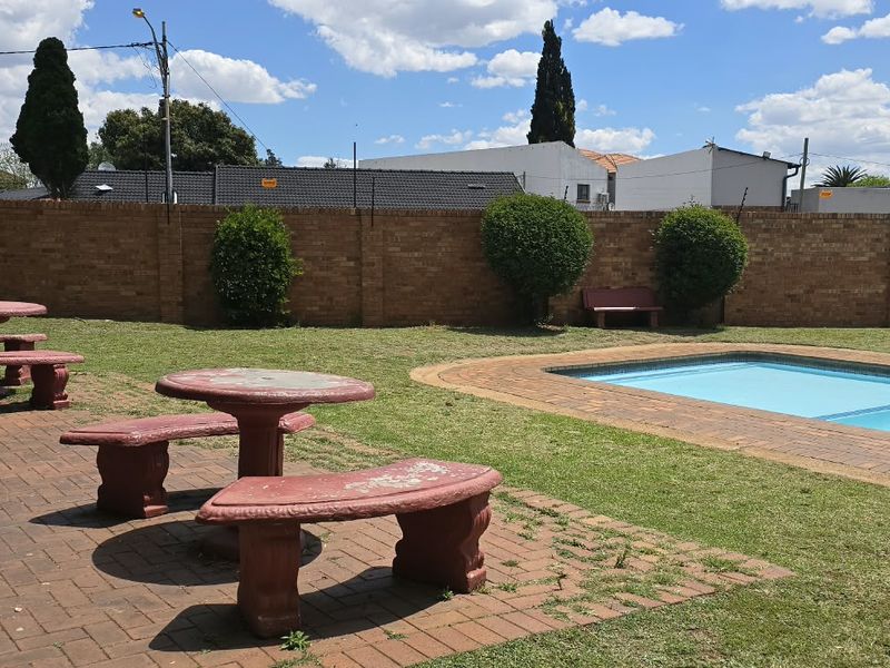 Communal pool area featuring a paved patio, swimming pool, and brick perimeter wall with electric fencing in a well-maintained garden setting.