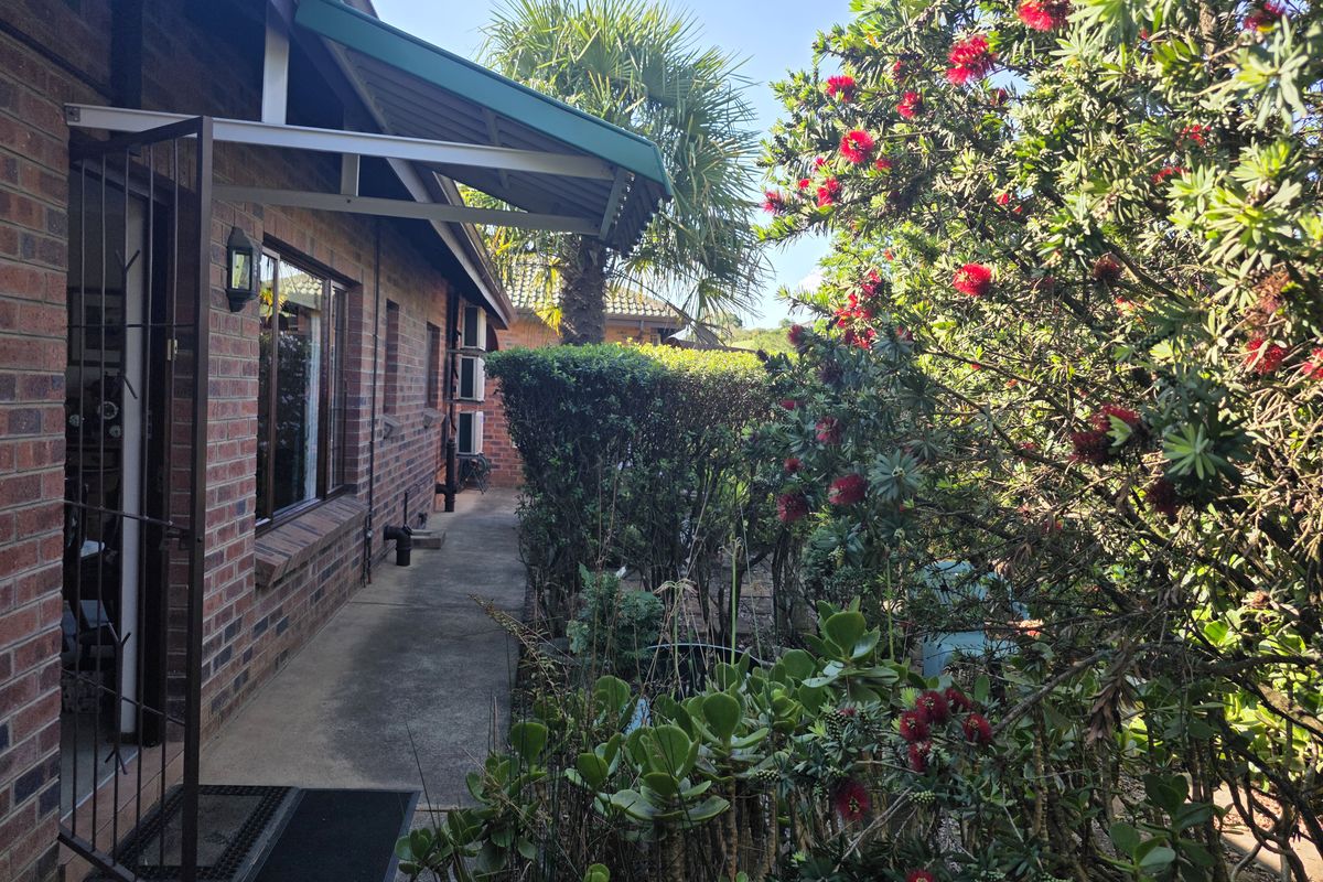 A well-maintained rear exterior view of the townhouse featuring a brick facade, concrete walkway, and established garden with a protective metal awning.