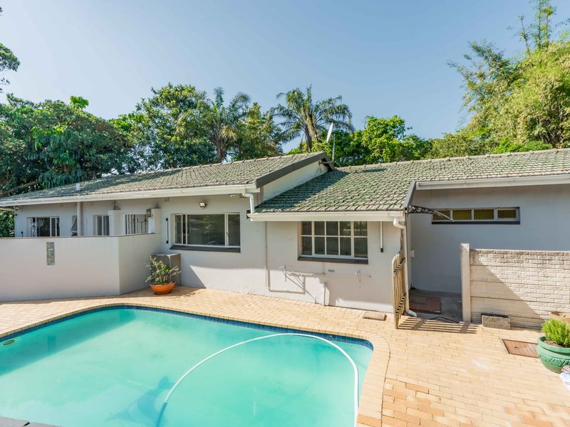 A rear exterior view featuring a swimming pool with brick paving, set against the house facade with a tiled roof and established garden surroundings.