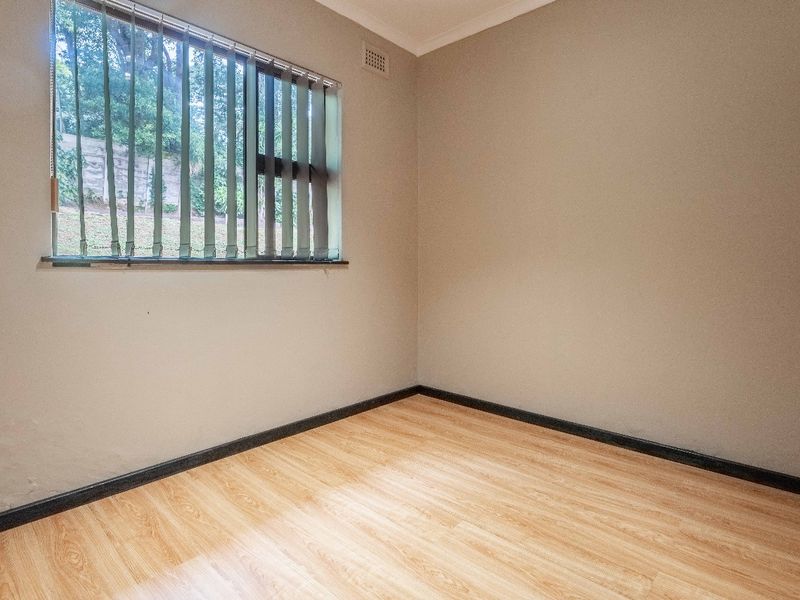 A clean, empty bedroom featuring light-toned laminate flooring, neutral walls, and vertical window blinds.