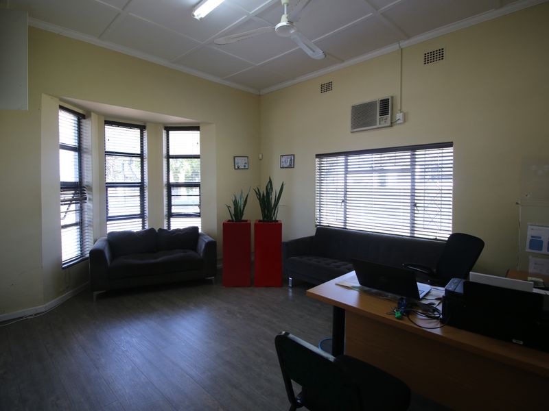 A functional office space featuring laminate flooring, a bay window with venetian blinds, a ceiling fan, and a wall-mounted air conditioning unit.