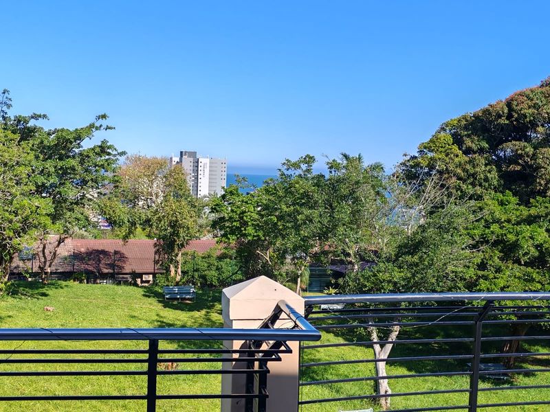 A scenic elevated view overlooking a lush green garden area with distant coastal buildings and ocean horizon visible in the background.