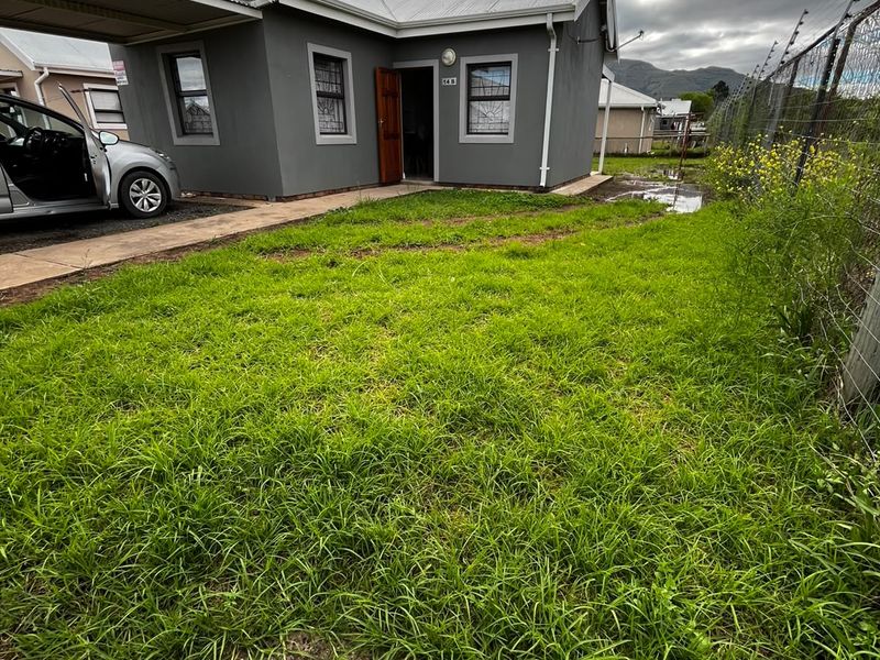 Front exterior view of a single-story house featuring a carport, solar water heater, and security fencing, set on a grassed lot.