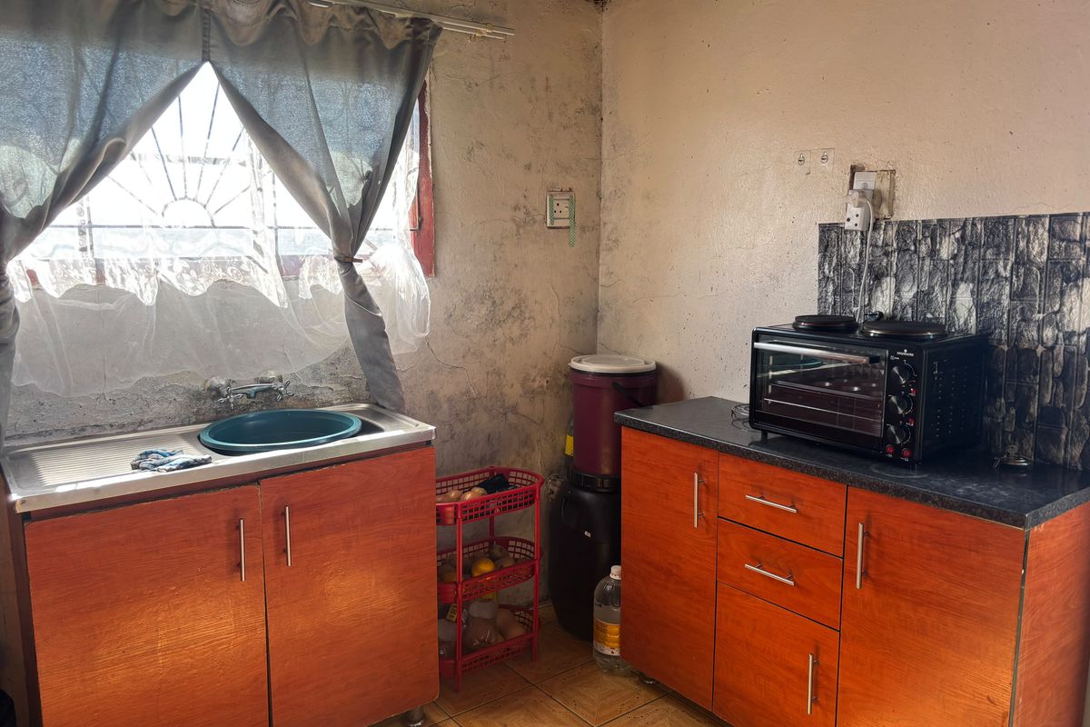 A compact kitchen area featuring wooden cabinetry, a stainless steel sink, and tiled flooring, currently requiring cosmetic updates and wall repairs.