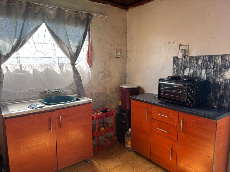 A compact kitchen area featuring wooden cabinetry, a stainless steel sink, and tiled flooring, currently requiring cosmetic updates and wall repairs.