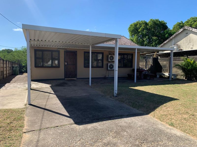 Rear exterior view of the house featuring a large metal carport, concrete driveway, and established garden area with perimeter walling.
