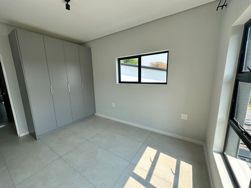 A bright, modern bedroom featuring neutral tiled flooring, built-in grey cupboards, and large aluminium-framed windows providing ample natural light.