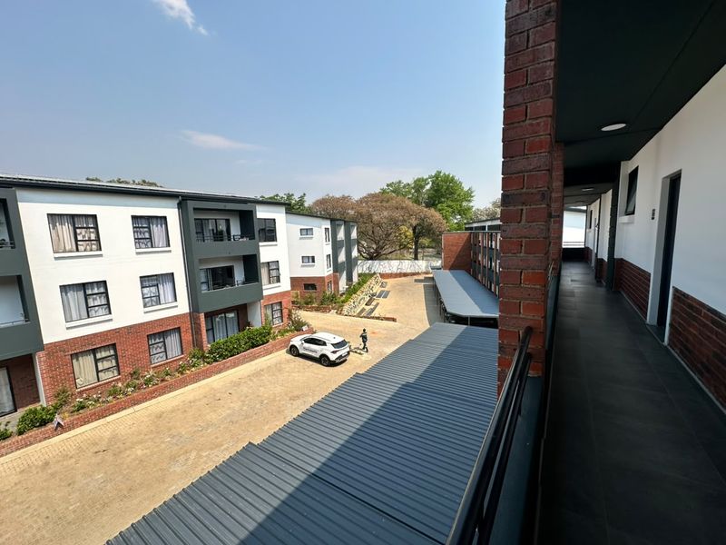 An elevated view of the apartment complex exterior, featuring a paved courtyard, covered parking bays, and a brick-accented building facade with a tiled walkway.