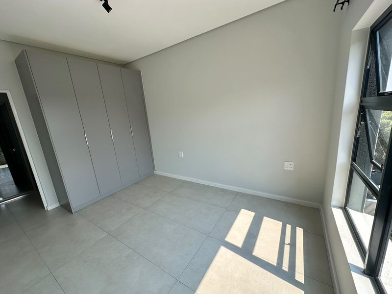 A bright, modern bedroom featuring neutral tiled flooring, built-in grey cupboards, and large aluminium-framed windows, offering a clean and contemporary living space.