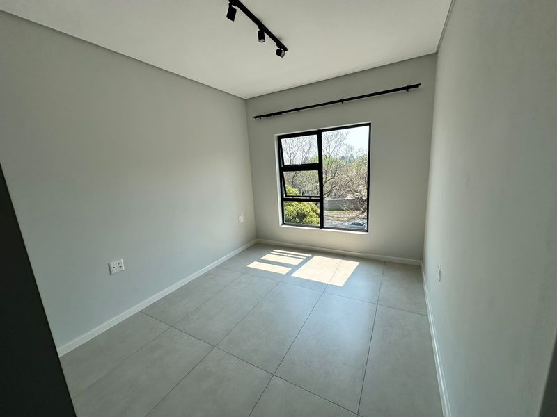 A bright, modern, and empty bedroom featuring neutral tiled flooring, black aluminium window frames, and contemporary track lighting.
