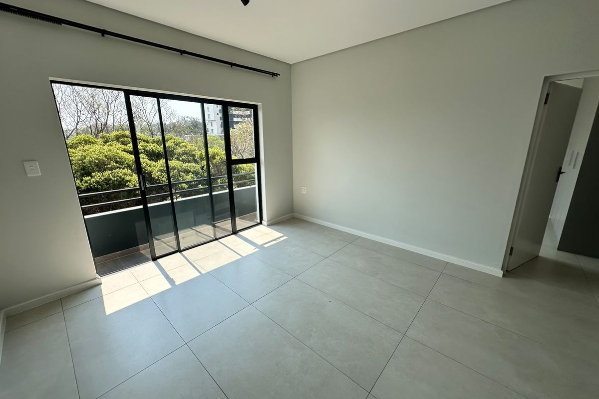 A bright, empty bedroom featuring large format floor tiles, modern track lighting, and a sliding glass door leading to a private balcony.