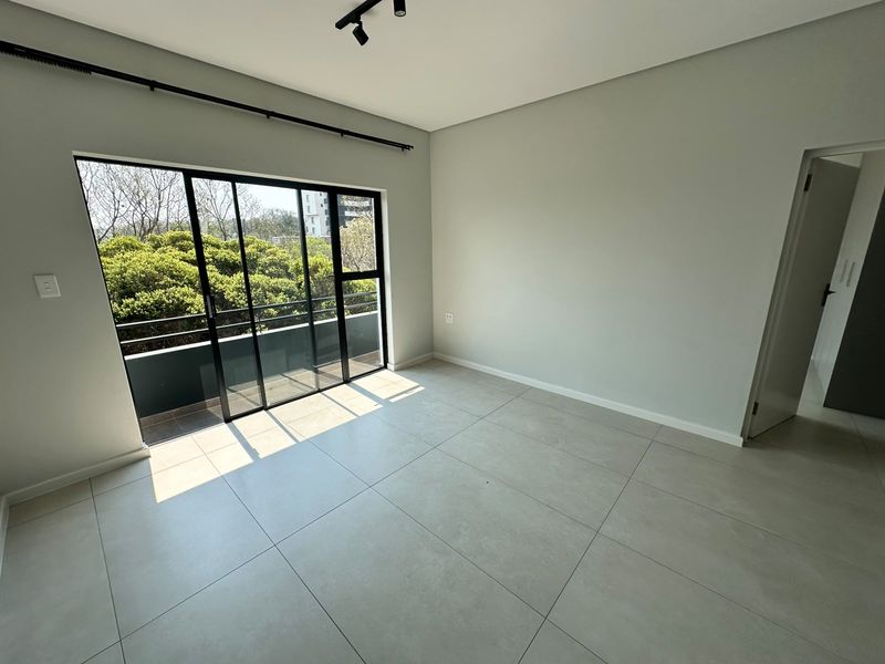 A bright, empty bedroom featuring large format floor tiles, modern track lighting, and a sliding glass door leading to a private balcony.