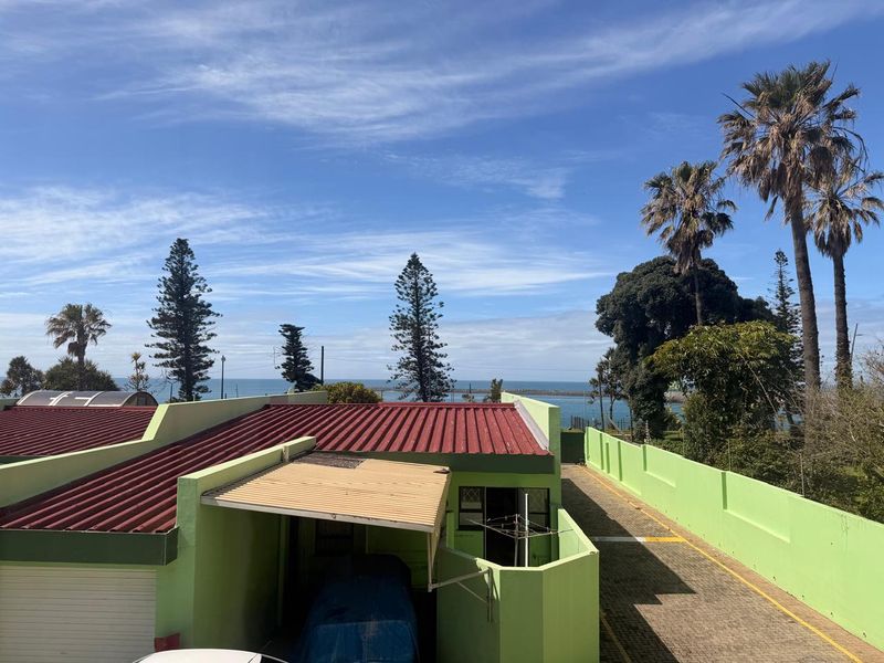 An elevated view of the rear exterior of the townhouse, featuring a paved driveway, carport, and boundary wall with a distant ocean view.