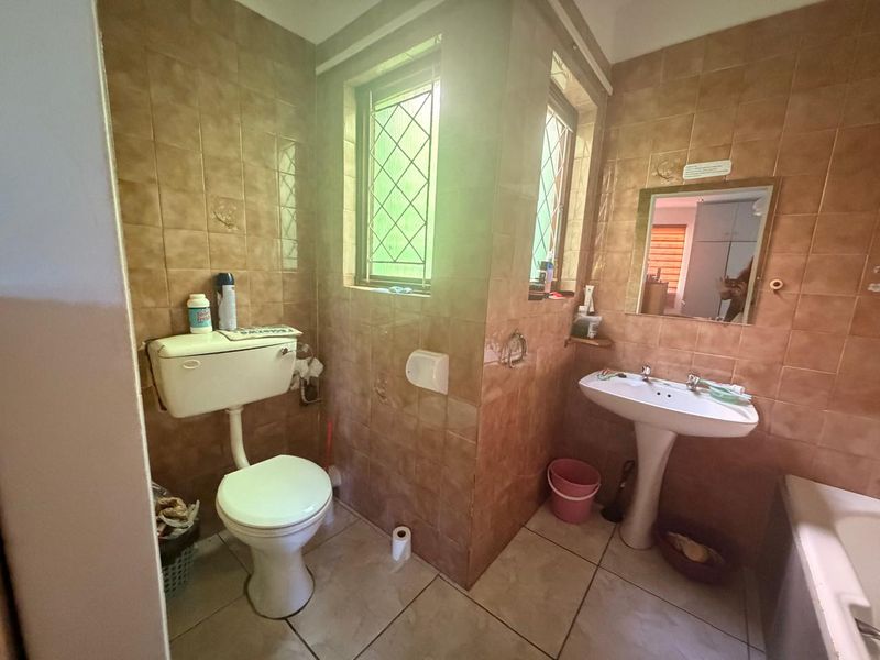 A dated bathroom featuring full-height wall tiling, a pedestal basin, and a standard toilet, with natural light provided by a window with security bars.