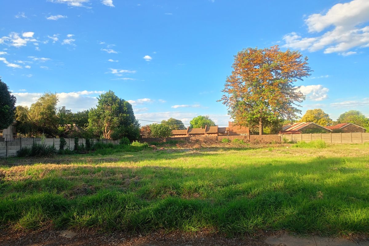 A vacant residential plot featuring established grass, mature trees, and a perimeter wall, offering a clear space for future development.