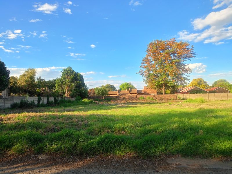 A vacant residential plot featuring established grass, mature trees, and a perimeter wall, offering a clear space for future development.