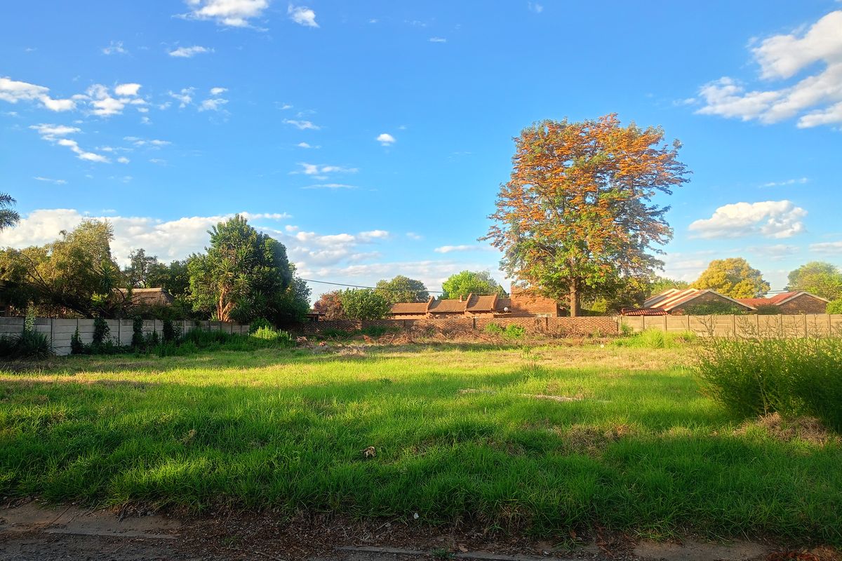 A vacant residential plot featuring established grass, mature trees, and a perimeter wall, offering a clear space for future development.