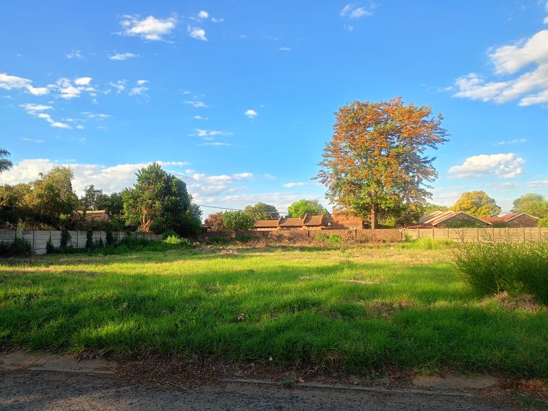 A vacant residential plot featuring established grass, mature trees, and a perimeter wall, offering a clear space for future development.