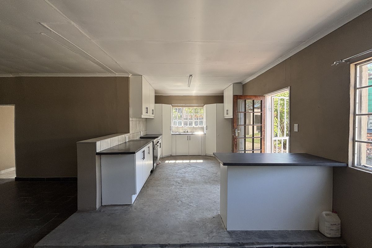 An empty, open-plan kitchen featuring white cabinetry, dark countertops, and direct access to the garden through a timber-framed door.