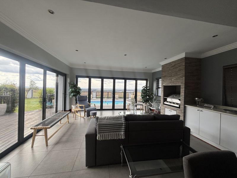 Spacious open-plan living area featuring a built-in braai, stone-clad fireplace, and large aluminium stacking doors opening onto a deck with pool views.