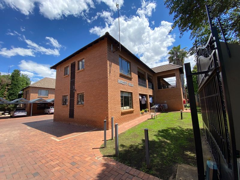 Exterior view of a multi-story face brick apartment complex featuring a paved driveway, covered parking bays, and well-maintained communal garden areas.