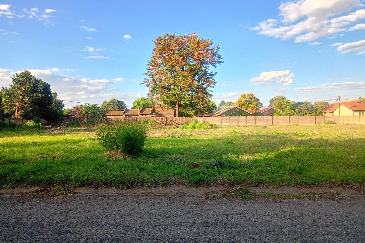 A vacant residential plot featuring a grassed surface, asphalt road frontage, and a precast concrete boundary wall at the rear.