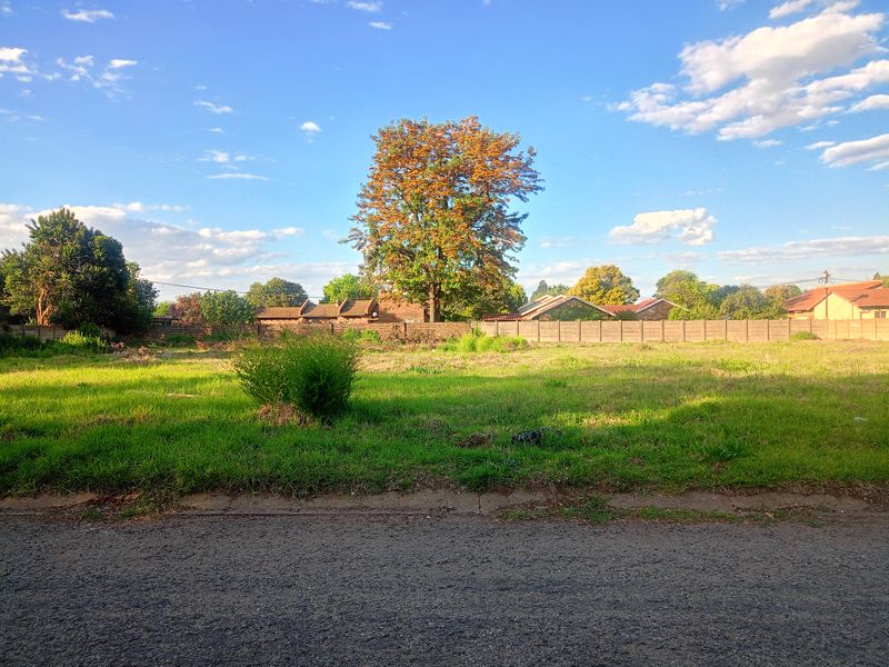 A vacant residential plot featuring a grassed surface, asphalt road frontage, and a precast concrete boundary wall at the rear.