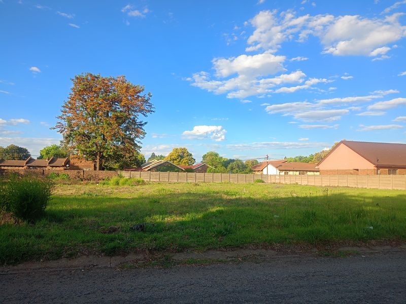 A vacant residential plot featuring a grassy surface, enclosed by a precast concrete boundary wall with a mature tree on the property.