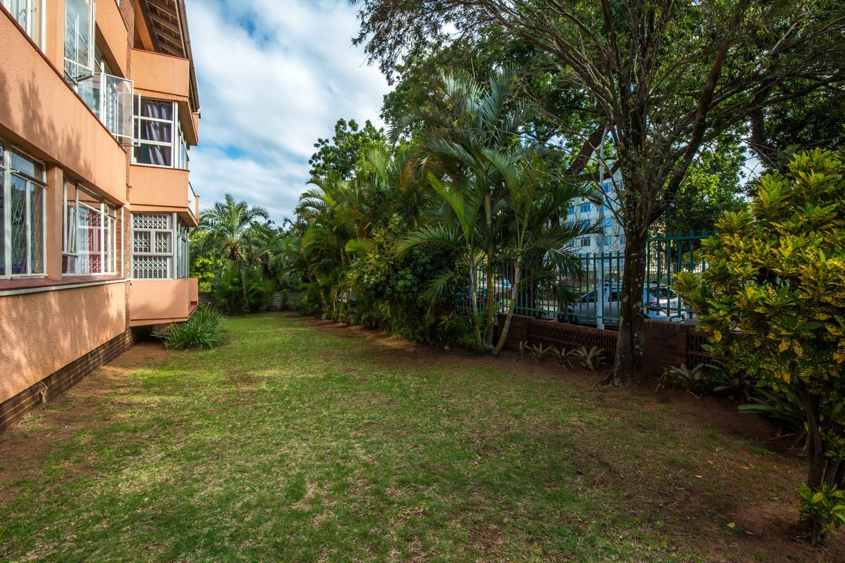 A view of the rear garden area featuring a lawn, mature trees, and a perimeter fence alongside the apartment building exterior.
