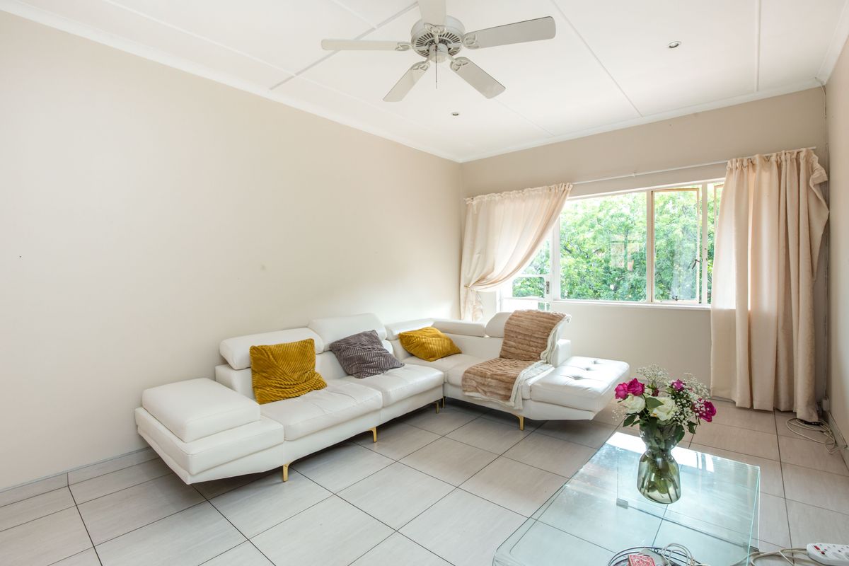 A bright living room featuring neutral tiled flooring, a ceiling fan, and a large window providing natural light and garden views.