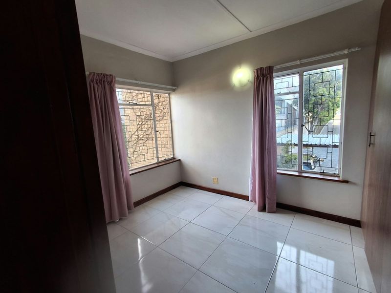 A bright, empty bedroom featuring tiled flooring, built-in cupboards, and large windows fitted with security bars.