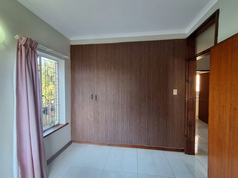 A bright, empty bedroom featuring tiled flooring, built-in wooden cupboards, and a window with security bars, offering a clean canvas for personalization.