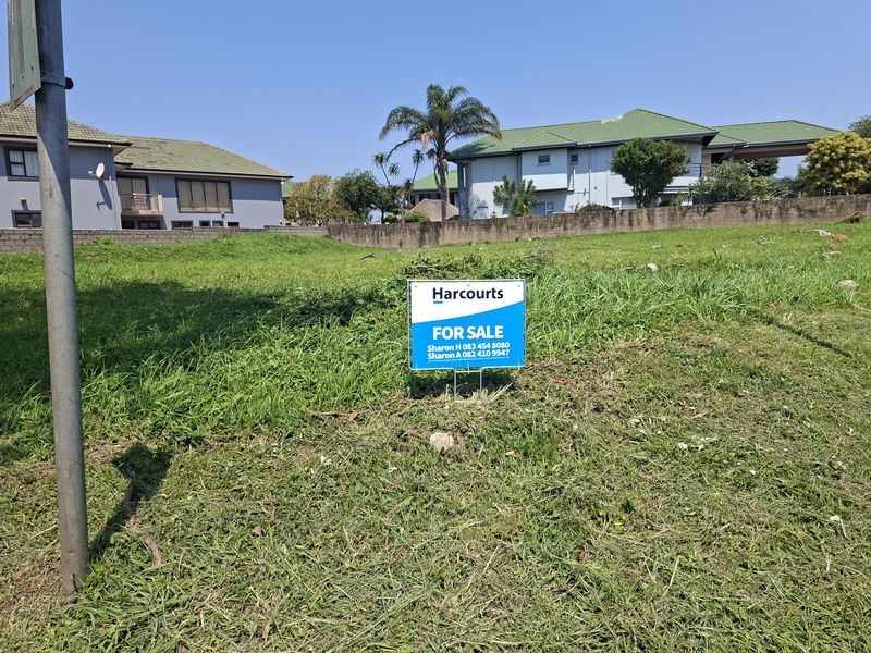 A vacant residential plot of land featuring a gentle slope and a retaining wall, currently marked with a real estate for sale sign.