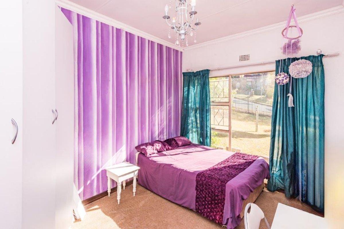 A bedroom featuring a built-in wardrobe, a decorative chandelier, and a large window with security bars, offering natural light and garden views.
