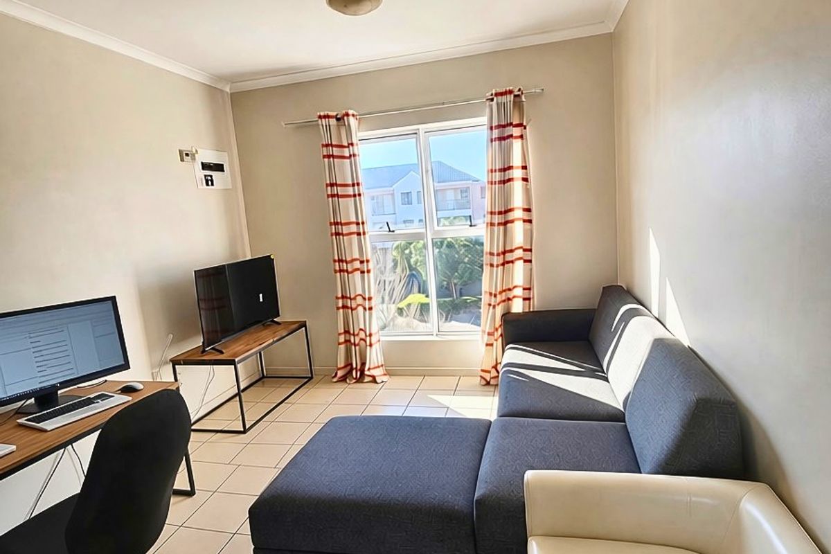 A functional living room featuring neutral tiled flooring, cove cornicing, and a large aluminium-framed window allowing for natural light.