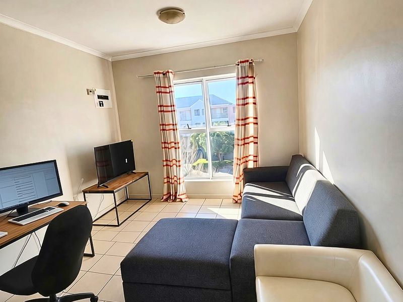 A functional living room featuring neutral tiled flooring, cove cornicing, and a large aluminium-framed window allowing for natural light.